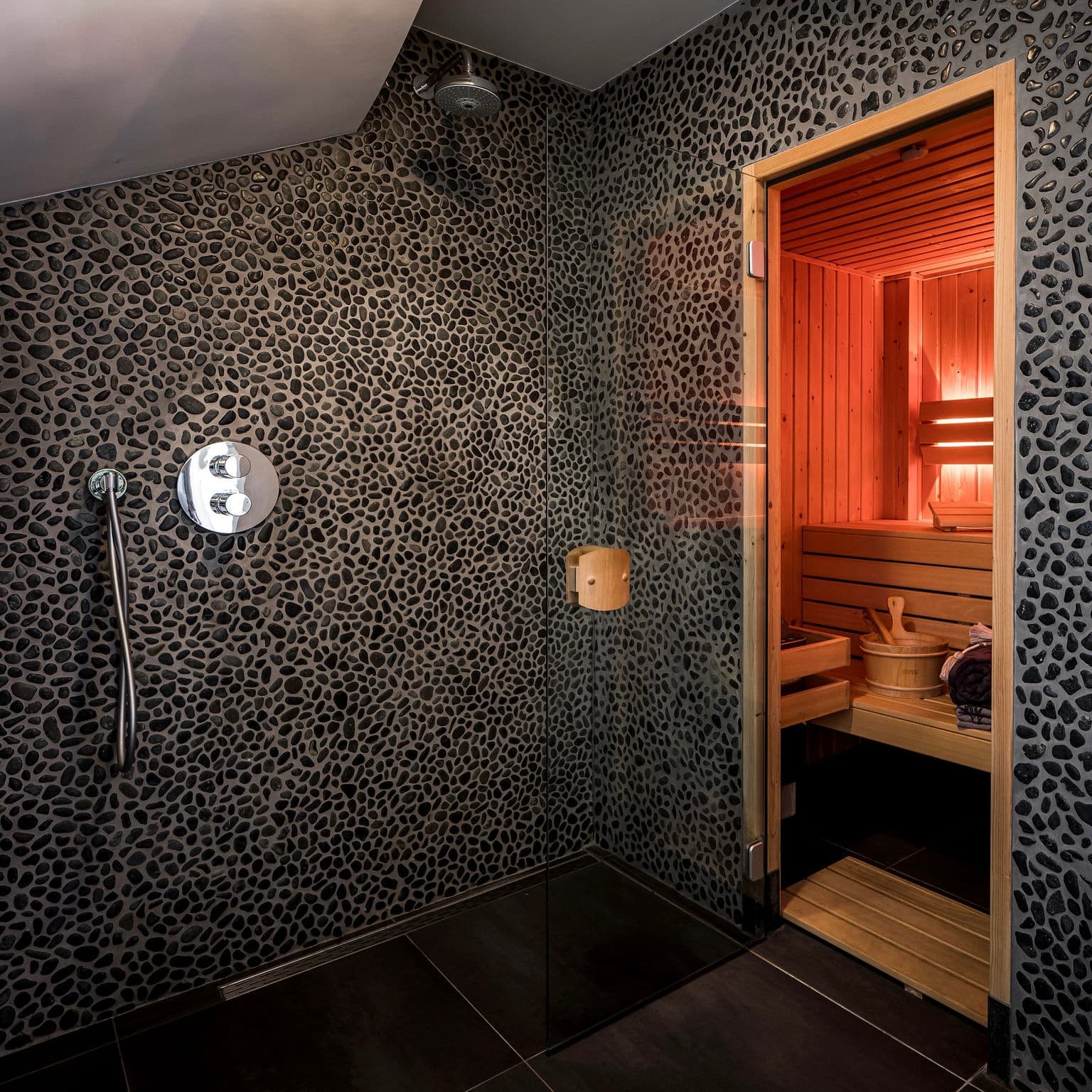 Begehbare Regendusche mit schwarzen Wänden im Kieselmuster und Blick in die private Holzsauna des Romantic Wellness B&B.