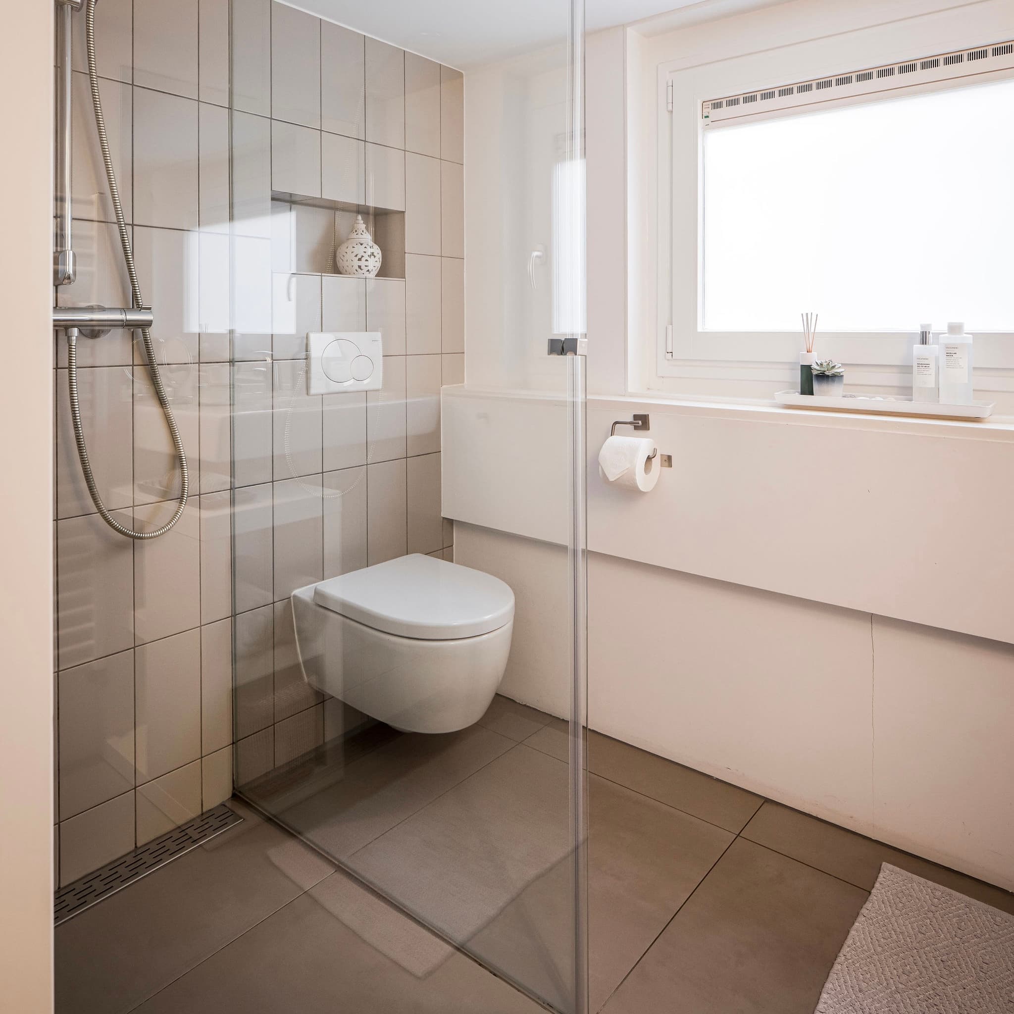 Modernes Badezimmer mit wandhängendem WC, Glasabtrennung, beigefarbenen Fliesenwänden, hellem Fenster und kleinem Teppich.