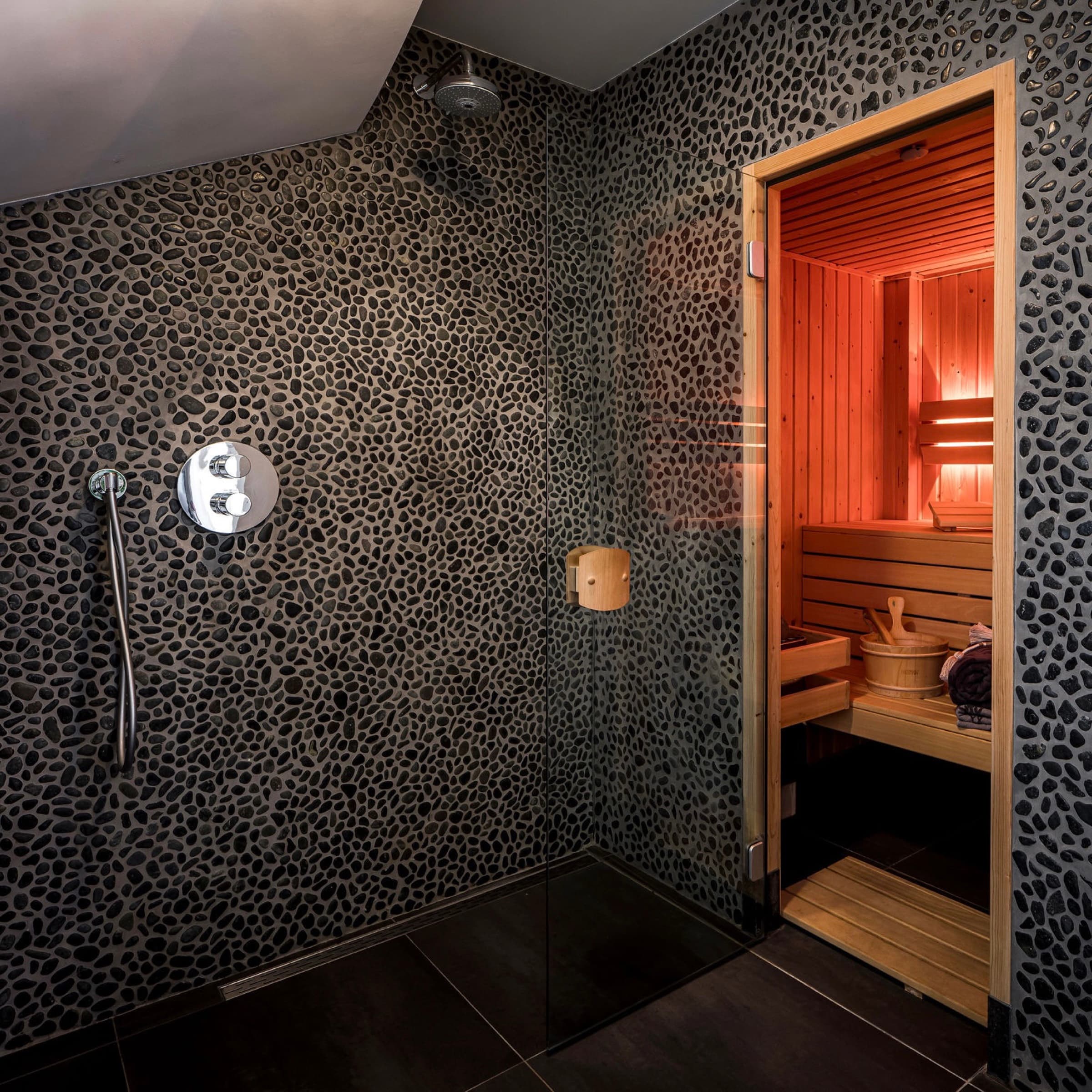 Ducha de lluvia a ras de suelo junto a la sauna finlandesa privada de Romantic Wellness B&B en Noordwijk, con paredes oscuras de diseño de guijarros.